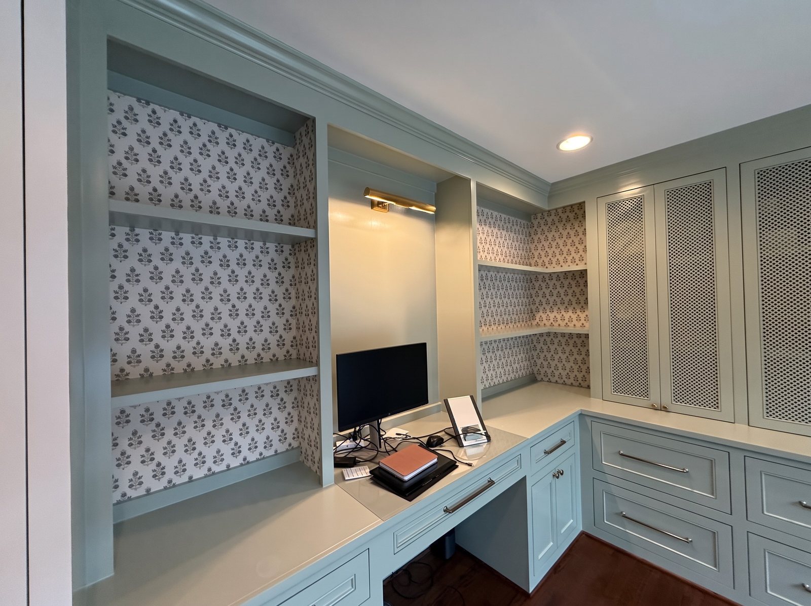 Pale blue-green built-in desk and shelving with a small floral block-print wallpaper on the wall behind the shelves.