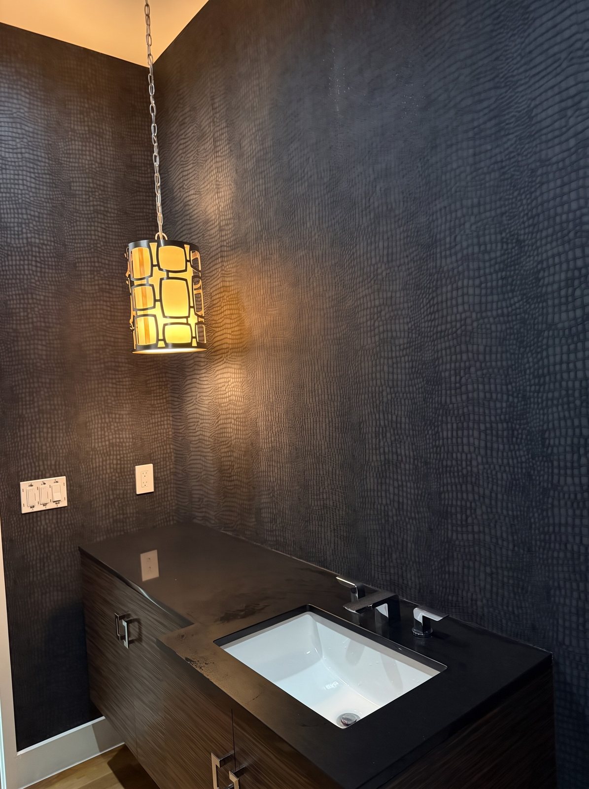 Dark navy crocodile-embossed textured wallcovering behind a dark vanity with a glowing pendant light.