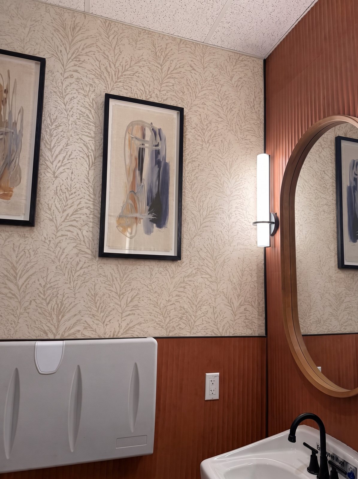 Commercial restroom with cream fern-pattern wallpaper above wood slat wainscot, an oval mirror and framed art.