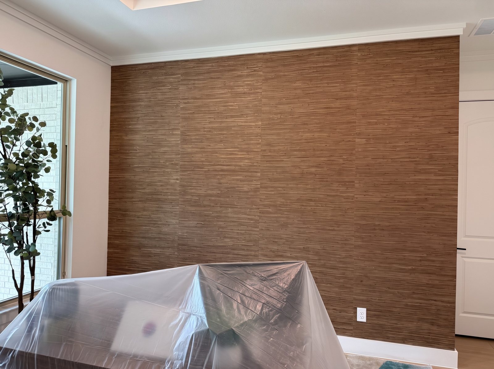 Wall covered in warm brown woven grasscloth with visible panel joins; furniture in front is protected with plastic sheeting.