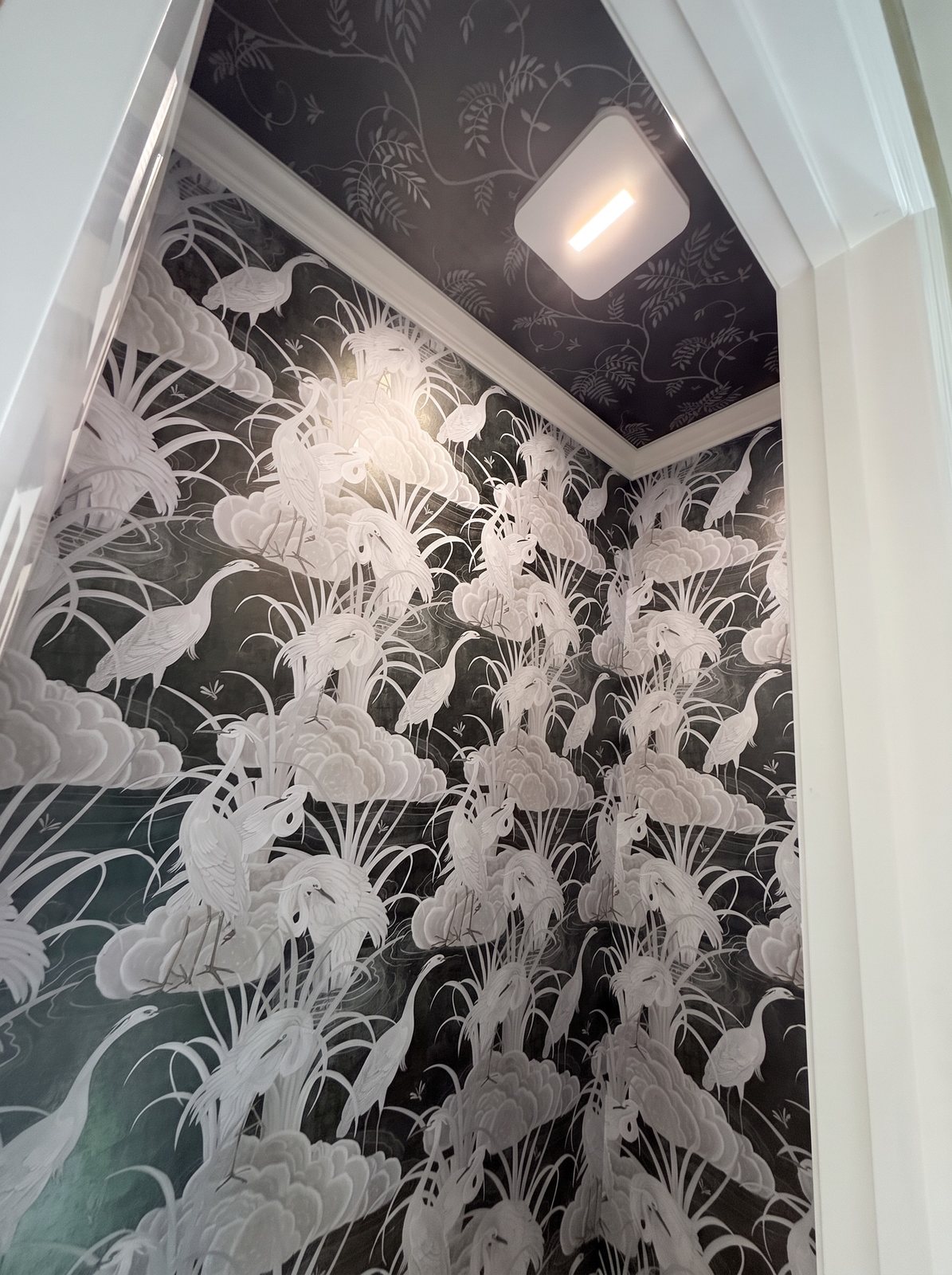 Powder room with black wallpaper of white herons and clouds on the walls and a dark floral paper on the ceiling.