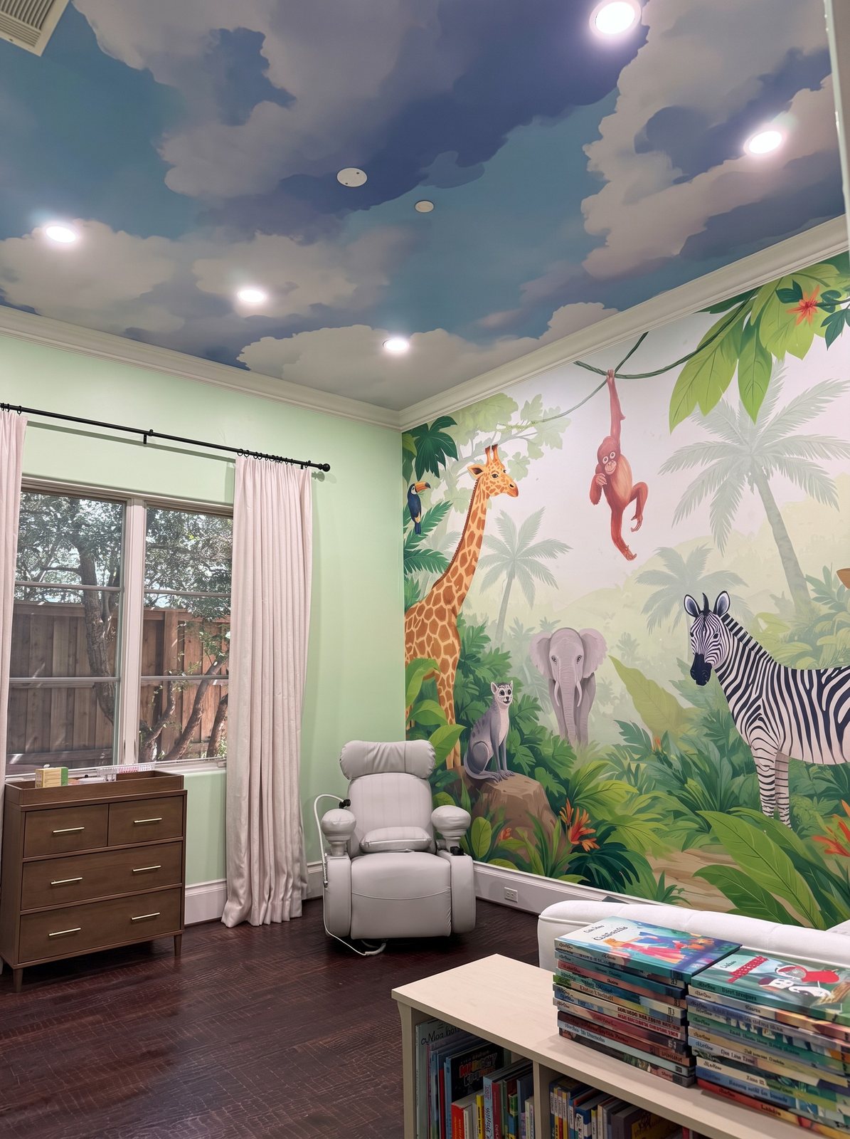 Nursery with a jungle mural of a giraffe, monkey, elephant and zebra on one wall and a blue cloud-sky mural covering the ceiling.