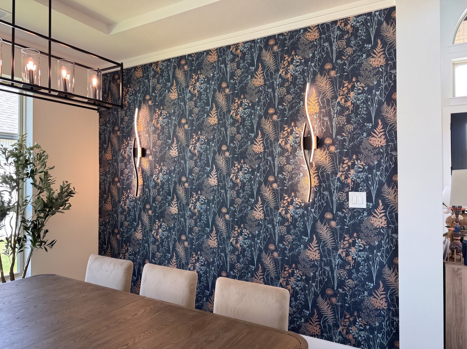 Dining room accent wall in navy wallpaper with copper and pale-blue ferns and wildflowers, two wave-shaped sconces on it.