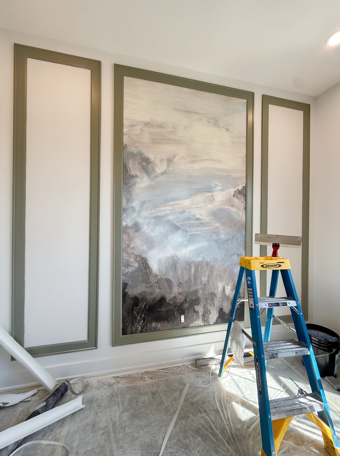 The framed mural wall during installation: centre panel finished, side panels still blank, a ladder and floor protection in the room.