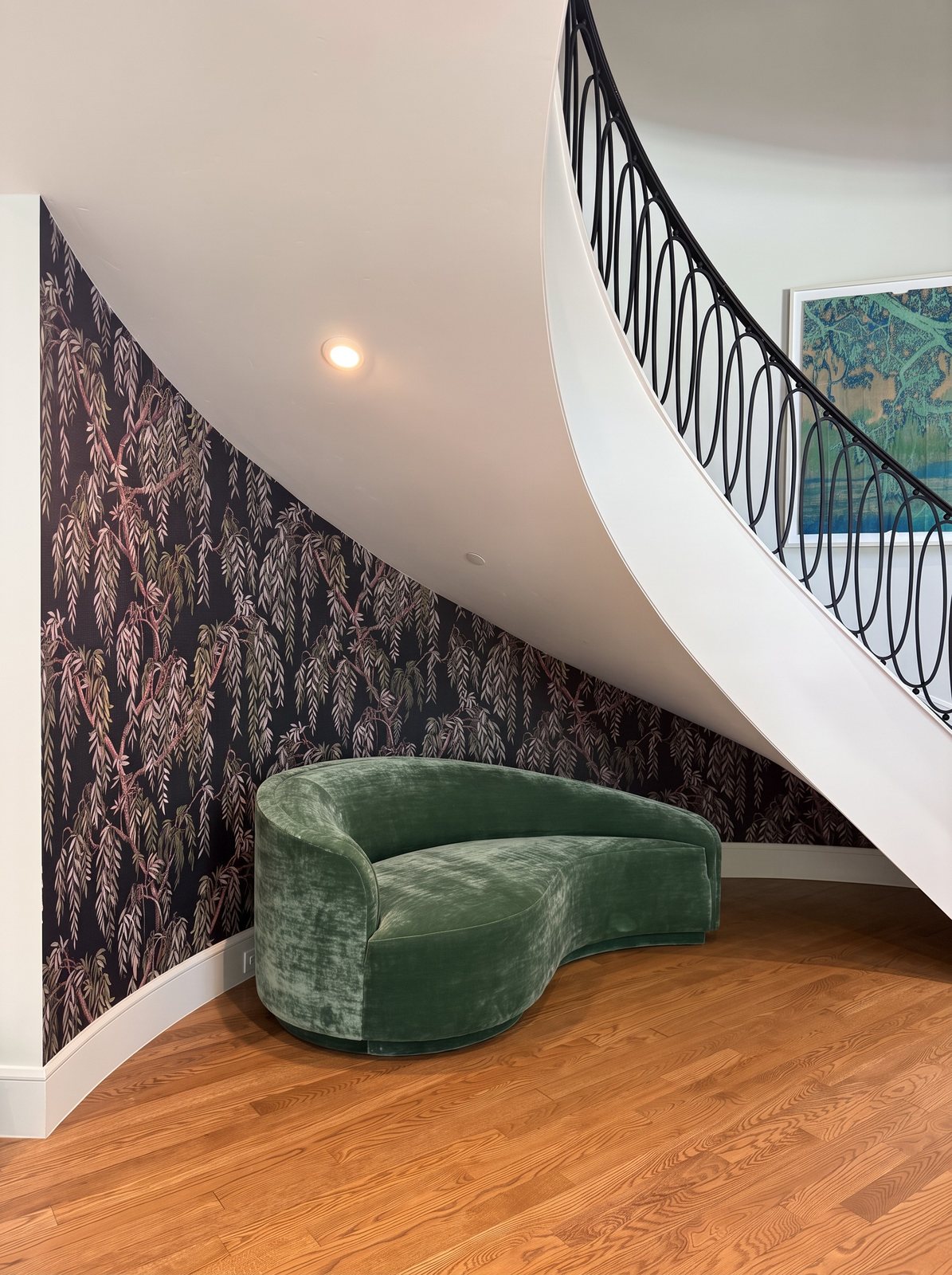 Curved wall beneath a sweeping staircase papered in a dark willow-branch pattern, with a green velvet sofa in front.